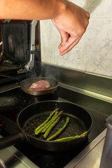 Two frying pans on an electric stove, one with green asparagus and the other with a duck breast, for a healthy gourmet meal.