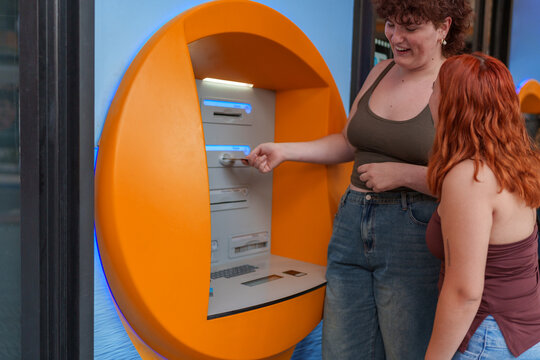 Young transgender woman using ATM with friend, managing finances and banking during daily routine - Powered by Adobe