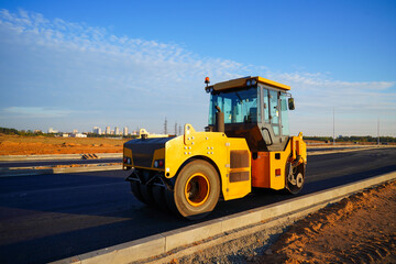 Obraz premium Heavy Machinery Compacts Asphalt on Construction Site. Road roller working on newly laid asphalt surface on sunset blue sky. Road Asphalting. Asphalt paving repair. Compactor machine on roadworks.