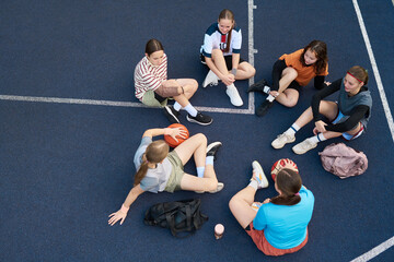 Group of six teenage girls sitting on outdoor basketball court resting after streetball game, holding basketballs and talking, diverse group including Caucasian and Black teenagers