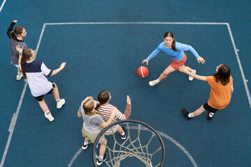 Group of multiethnic teenagers playing streetball on outdoor court, Caucasian teenage girl dribbling basketball while being guarded by Black teenage girl, teammates watching and preparing to assist