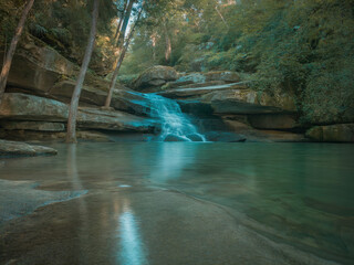 Beautiful tropical waterfall flowing into turquoise lagoon surrounded by lush green forest, peaceful nature landscape with crystal clear water, rocks and sunlight reflections in jungle