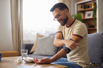 A man sits on a sofa in a well-lit living room, rolling up his sleeve to check his blood pressure using a monitor on the table beside him. A glass of water sits nearby.