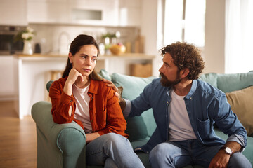 Fototapeta premium Two adults sit on a couch, deep in conversation about their relationship issues. The woman looks concerned while the man offers comfort in a bright and inviting living room.