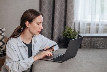 Focused woman uses her smartphone while sitting at a laptop, engaged in business communication or chatting.