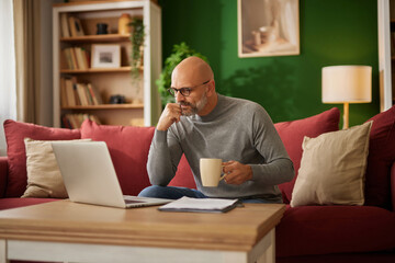 Fototapeta premium A man sits comfortably on a red couch, sipping coffee and analyzing financial documents on his laptop in a warm, inviting living room environment.