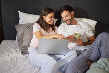 Fototapeta premium A couple is happily exploring vacation destinations on a laptop while lounging comfortably in bed, sharing ideas and sipping drinks during their cozy planning session.