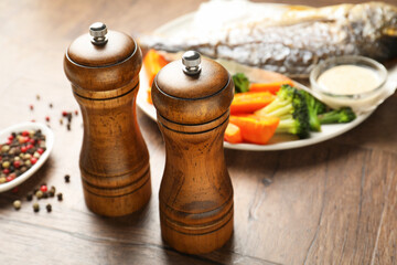 Salt, pepper mills, tasty roasted dorado fish, vegetables, sauce and peppercorns on wooden table,...