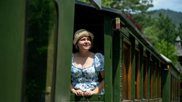 A joyful, young Caucasian woman in a sunhat revels in a scenic train journey, echoing Wanderlust Day vibes