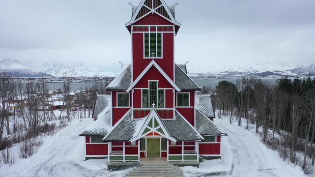 Aerial view of the striking red Buksnes Church against the stark white snow, contrasting with the distant mountains, Gravdal, Nordland, Norway.