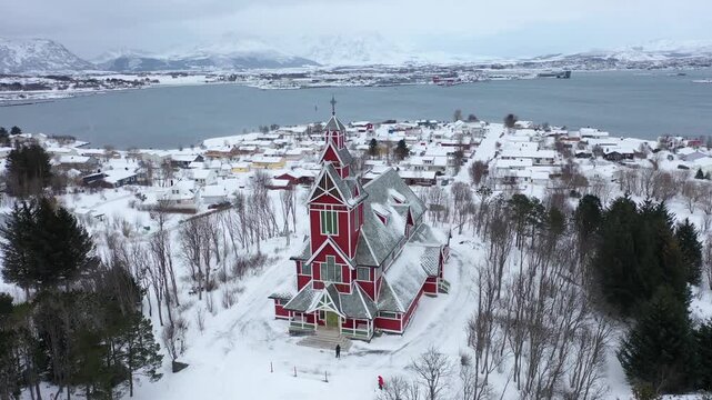 Aerial view of the striking red Buksnes Church amidst a snowy landscape, creating a beautiful contrast with the surrounding white and grey tones, Gravdal, Nordland, Norway.