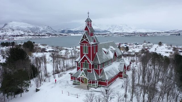 Aerial view of the striking red Buksnes Church amidst a snow-covered landscape with mountains and water in the background, Gravdal, Nordland, Norway.