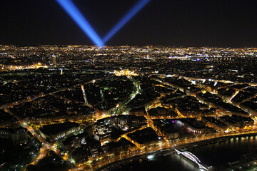 A bird's-eye view of Paris at night: bright street lights, the city skyline, and the bright blue beams of spotlights crossing the sky. 