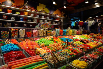A vibrant display of colorful bulk candies, gummy sweets, and lollipops at a market stall in the famous La Boqueria in Barcelona, Spain.