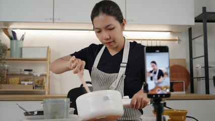 Asian woman in apron stirs ingredients while streaming a cooking vlog at home kitchen, smiling with confidence as an online influencer sharing recipes.