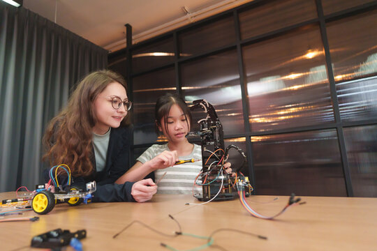 Asian girl practicing robotics with caucasian teacher support using laptop and robot model on wooden table.