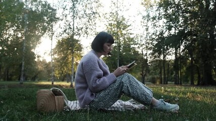 Young woman reading ebook sitting on grass in public park. Concept of recreating, resting, mindfulness, leisure without gadgets. Young woman is focused on reading or studying. - Powered by Adobe