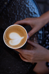 Hand holding a cup of coffee with heart-shaped foam design on a textured table outdoors in a cozy setting