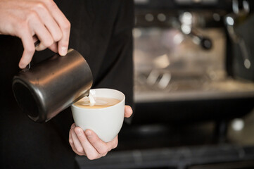 Barista pouring steamed milk into espresso to create latte art in a café setting. Close-up of hands, cup, and pitcher with blurred coffee machine in background