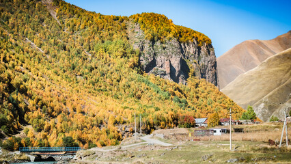 Colorful autumn forest near Truso Gorge in Georgia with a rural village, mountain cliffs and a small blue bridge under clear blue sky. Tranquil and scenic highland landscape