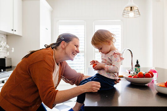 Mother holding toddler daughter seated on countertop holding an orange