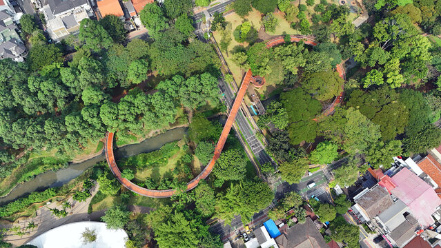 Aerial view of a vibrant orange pedestrian bridge winding gracefully through a lush green urban park, harmonizing nature and architecture, Jakarta, Indonesia.