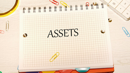 On a desk, a notebook displays the word ASSETS. Paperclips, pushpins, and a calculator surround the notebook, creating a scene focused on financial planning