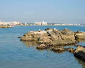 Low-angle view of a rugged, man-made rock breakwater in the calm sea. The modern skyline of the Italian resort town of Cattolica is visible in the background under a clear blue sky.