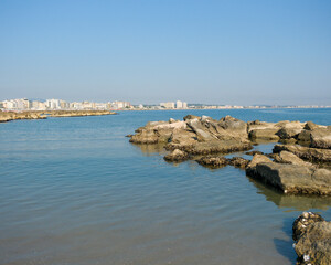 Low-angle view of a rugged, man-made rock breakwater in the calm sea. The modern skyline of the Italian resort town of Cattolica is visible in the background under a clear blue sky.