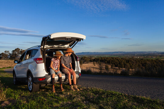 Teen boy and preteen girl siblings sitting in car boot beside rural Australian road in warm sunlight