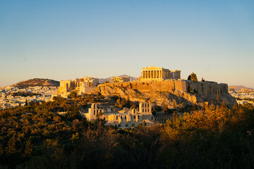 Fototapeta premium Acropolis of Athens during golden hour, with the Parthenon temple glowing above the city skyline. The soft evening light highlights the ancient ruins and the surrounding greenery, architecture