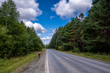 A woman walks along a highway with a forest on a summer day.