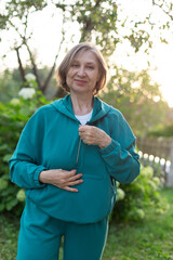 Happy smiling active mature woman zipping up blue sportswear hoodie outdoors. Sport training concept