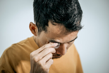 An Asian man is applying eye drops to himself. One hand holds the bottle above his eye, while the other pulls down his lower eyelid. The background is plain white.