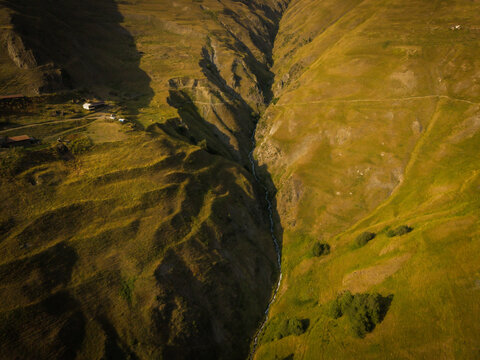 Aerial view of a stark, deep gorge cutting through verdant hills, shadows playing against the sun-kissed slopes, a lone settlement clinging to the distant edge, Dartlo, Kakheti, Georgia.