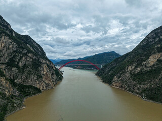 A large red bridge spans the wide river between steep green mountains under a cloudy sky in China's Three Gorges region.