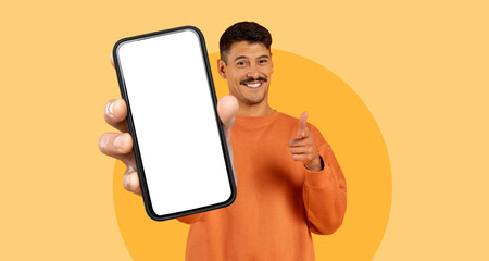A man stands cheerfully, pointing at the camera while holding a smartphone in front of him. He is dressed in a cozy sweater and his friendly expression enhances the casual vibe of the moment.