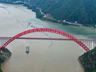 A red bridge spans a wide river, with a boat passing underneath its arches in Yichang Three Gorges,...