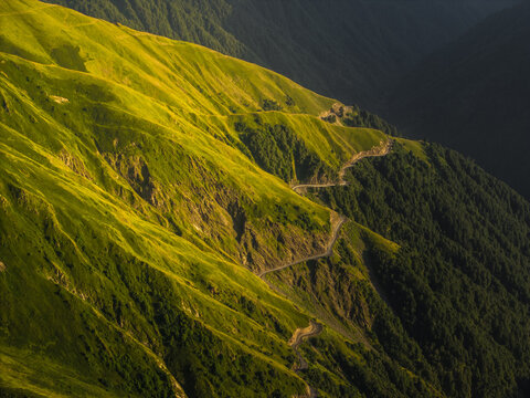 Aerial view of a winding road snakes through vibrant green mountains contrasting with deep shadows, Alazani pass, Chala, Kakheti, Georgia.
