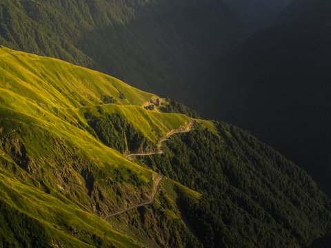 Aerial view of a winding road cutting through verdant mountainsides, the sun painting the slopes with gold against the deep shadows, Chala, Kakheti, Georgia.