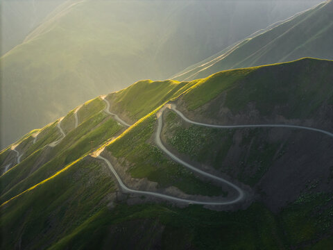 Aerial view of a serpentine road winding through verdant mountains under a soft, golden sunlight, Alazani pass, Chala, Kakheti, Georgia.