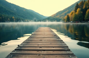 Obraz premium A wooden dock extends into a calm lake surrounded by forested mountains under a soft, misty sky