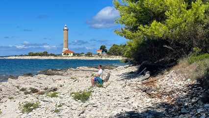 Man sitting on rocky shore facing a coastal lighthouse, surrounded by turquoise sea and pine trees under clear summer sky, symbolizing solitude and connection with nature