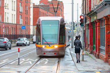 Naklejka premium A well-dressed stylish woman is walking on the sidewalk next to traffic lights - Public tram on the street in Dublin, İreland