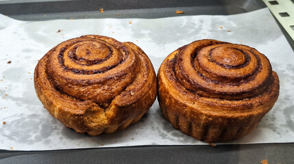 two cinnamon rolls on white parchment paper with the remaining crumbs beside it for sale. 