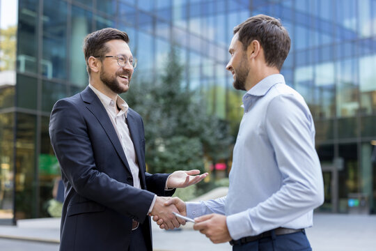 Two smiling professional businessmen are shaking hands, confirming a successful business deal or partnership in an urban setting with a modern office building in the background - Powered by Adobe