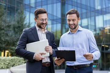 Two smiling adult businessmen, one holding a laptop and the other reviewing documents on a clipboard, are standing and discussing business matters outdoors in front of a modern corporate building