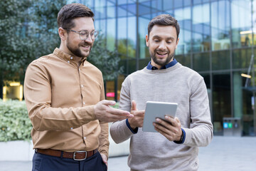 Two smiling adult businessmen engaged in a dynamic discussion, sharing ideas and collaborating on a project using a digital tablet outside a modern office building