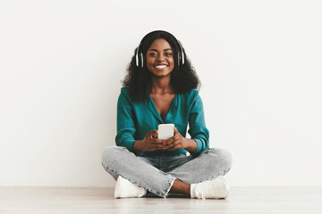 Cheerful young black woman in casual sitting on floor over white studio background, using modern...