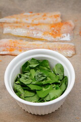Fresh fish fillets seasoned with spices on parchment paper, ready for cooking, next to a white ramekin bowl filled with vibrant green leaves, illustrating healthy meal preparation